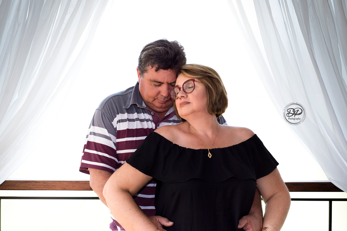 Casal juntos em uma sacada com cortinas brancas, em ensaio família feito em Bariri SP