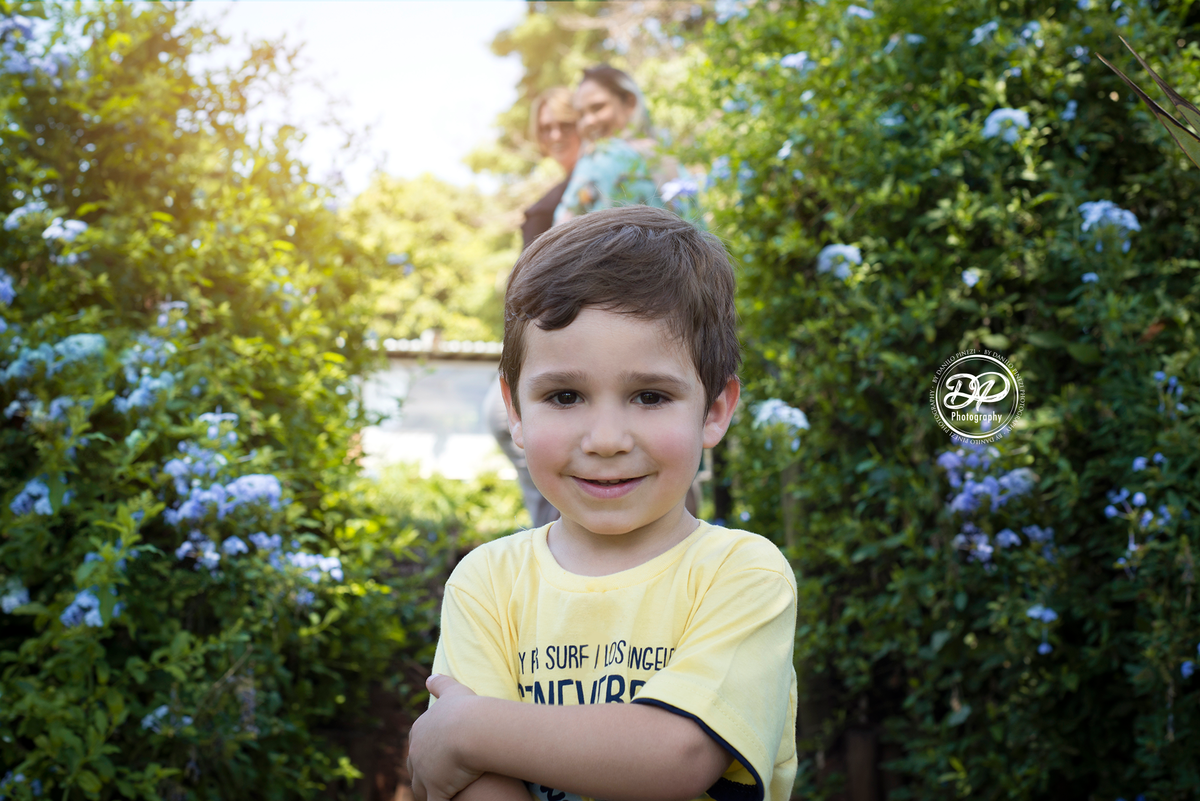 menino de braços cruzados em um jardim, com sua mamãe e vovó ao fundo.