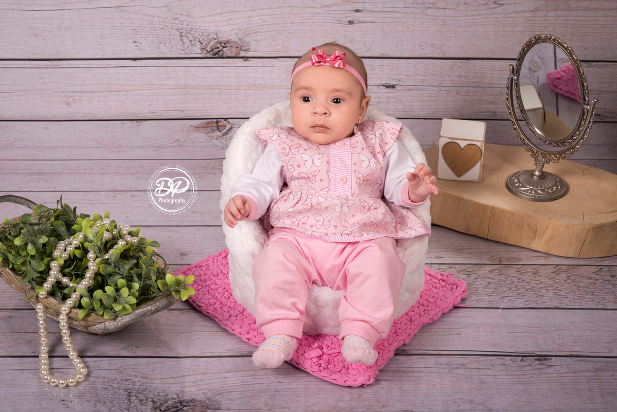 Sessão de 1 mês de bebê menina sentada em poltrona posicionadora com elementos como corações, colar de pérolas, folhagens e espero antigo, em sessão realizada no estúdio Danilo Pinezi Fotografia