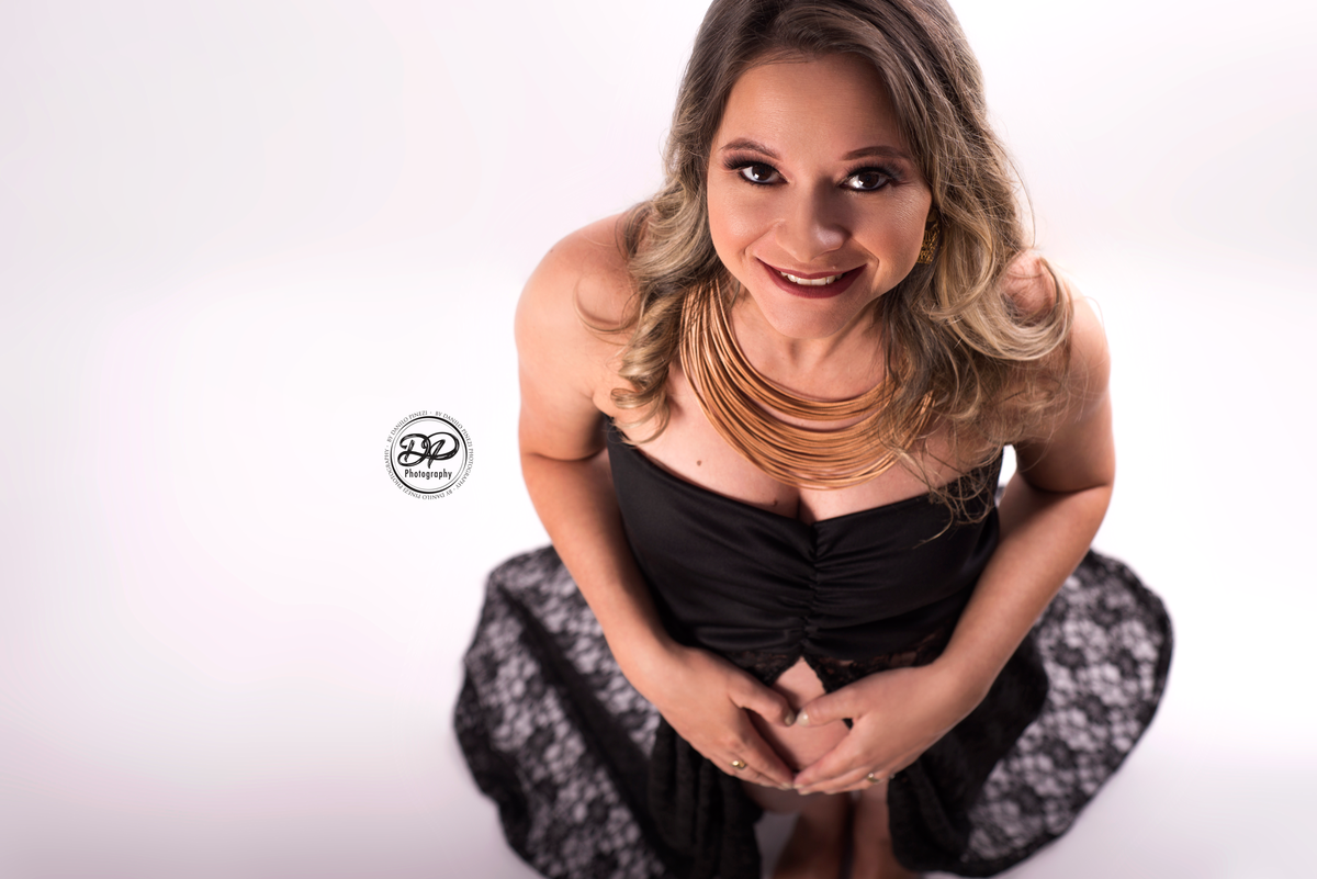Ensaio gestante com vestido de renda preto, olhando para cima, e fazendo um coração com as mãos em ensaio realizado no estúdio Danilo Pinezi Fotografia em Bariri.