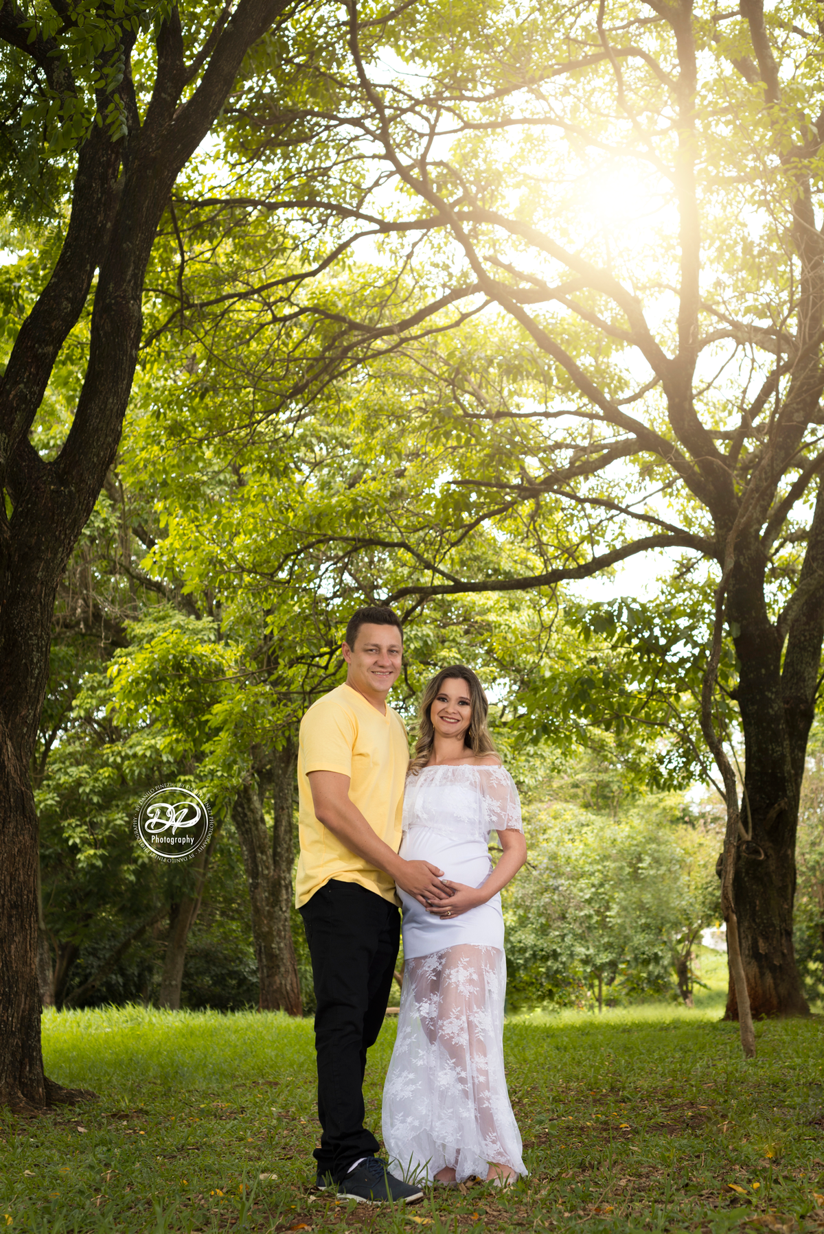 Ensaio gestante externo, gestante com vestido branco de renda, fotografia gestante casal, com copas das árvores e sol de fundo, executado pelo fotógrafo Danilo Pinezi em Bariri/SP