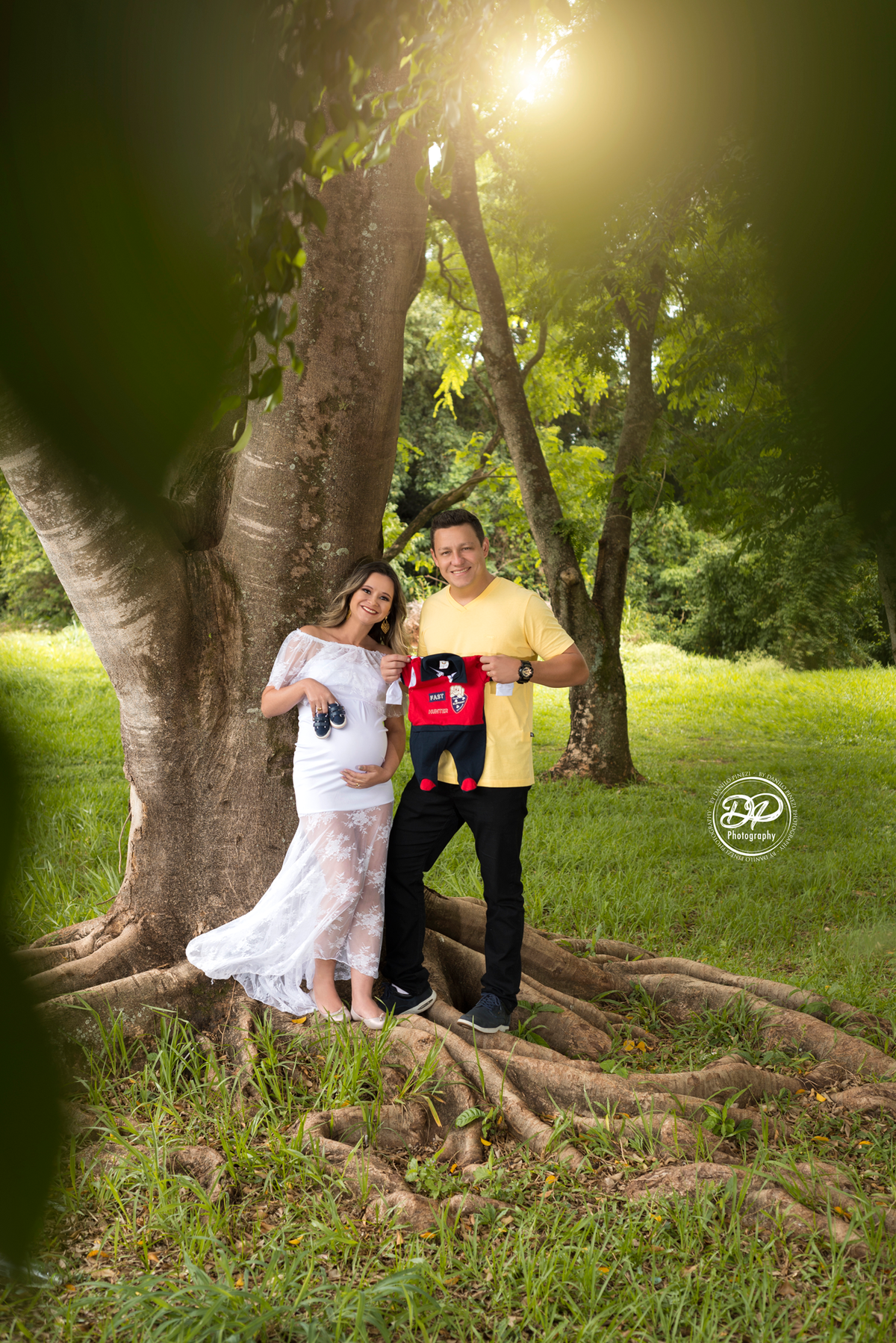 Ensaio gestante externo, gestante com vestido branco de renda, e papai de amarelo, segurando sapatinhos e roupinha do bebê fotografia gestante casal, executado pelo fotógrafo Danilo Pinezi em Bariri/SP