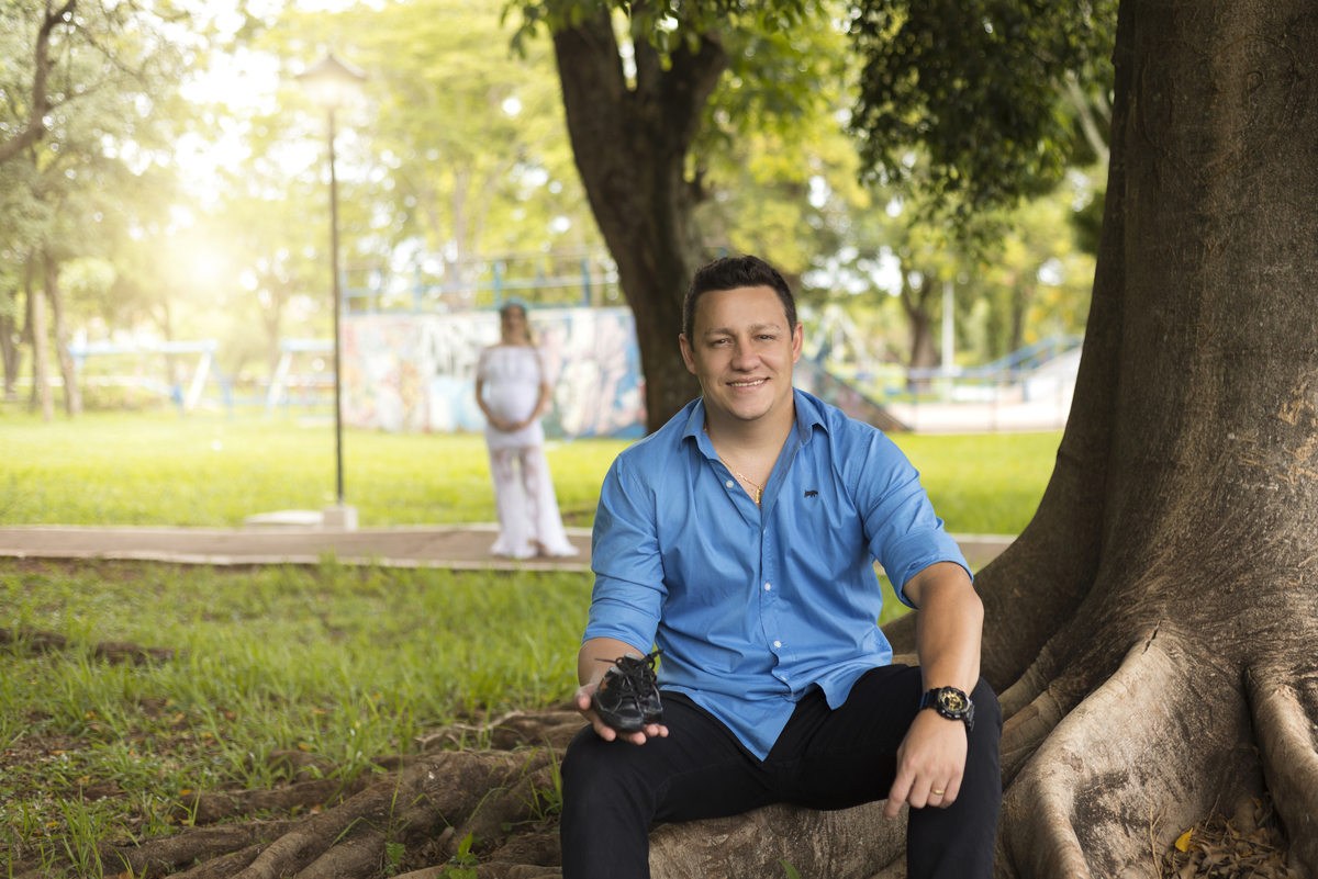 Ensaio gestante, papai sentado segurando um sapatinho de bebê e sorrindo, ao fundo a mamãe contra o sol, de vestido branco de renda e coroa de flores, executado pelo fotógrafo Danilo Pinezi em Bariri/SP