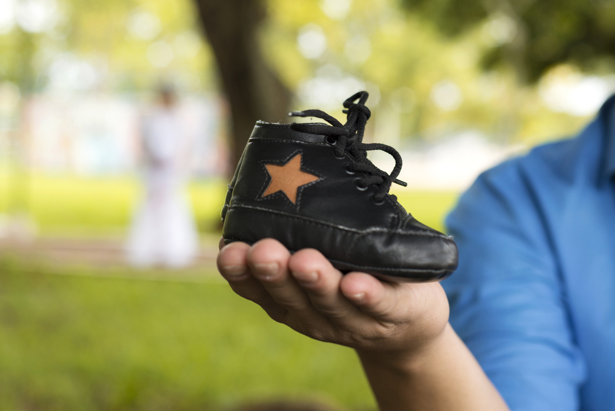 Ensaio gestante, a mão do papai em destaque  segurando um sapatinho de bebê preto com estrela marrom, ao fundo a mamãe contra o sol, de vestido branco de renda e coroa de flores, executado pelo fotógrafo Danilo Pinezi em Bariri/SP