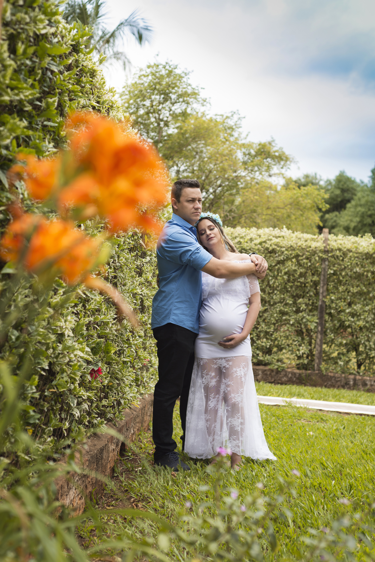 Ensaio gestante, casal abraçados em frente ao muro de plantas e e flores, mamãe de vestido branco de renda e coroa de flores, executado pelo fotógrafo Danilo Pinezi em Bariri/SP