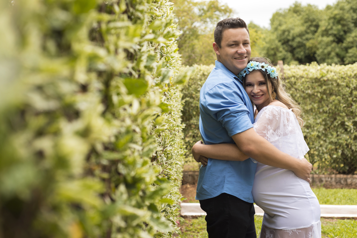 Ensaio gestante, casal abraçados em frente ao muro de plantas e e flores, mamãe de vestido branco de renda e coroa de flores, executado pelo fotógrafo Danilo Pinezi em Bariri/SP