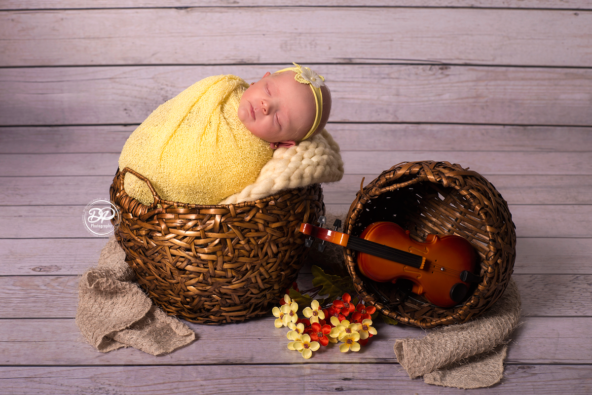 Ensaio newborn com a bebê enrolada no wrap amarelo posicionada dentro de um cesto, ornamentado com flores e um violino