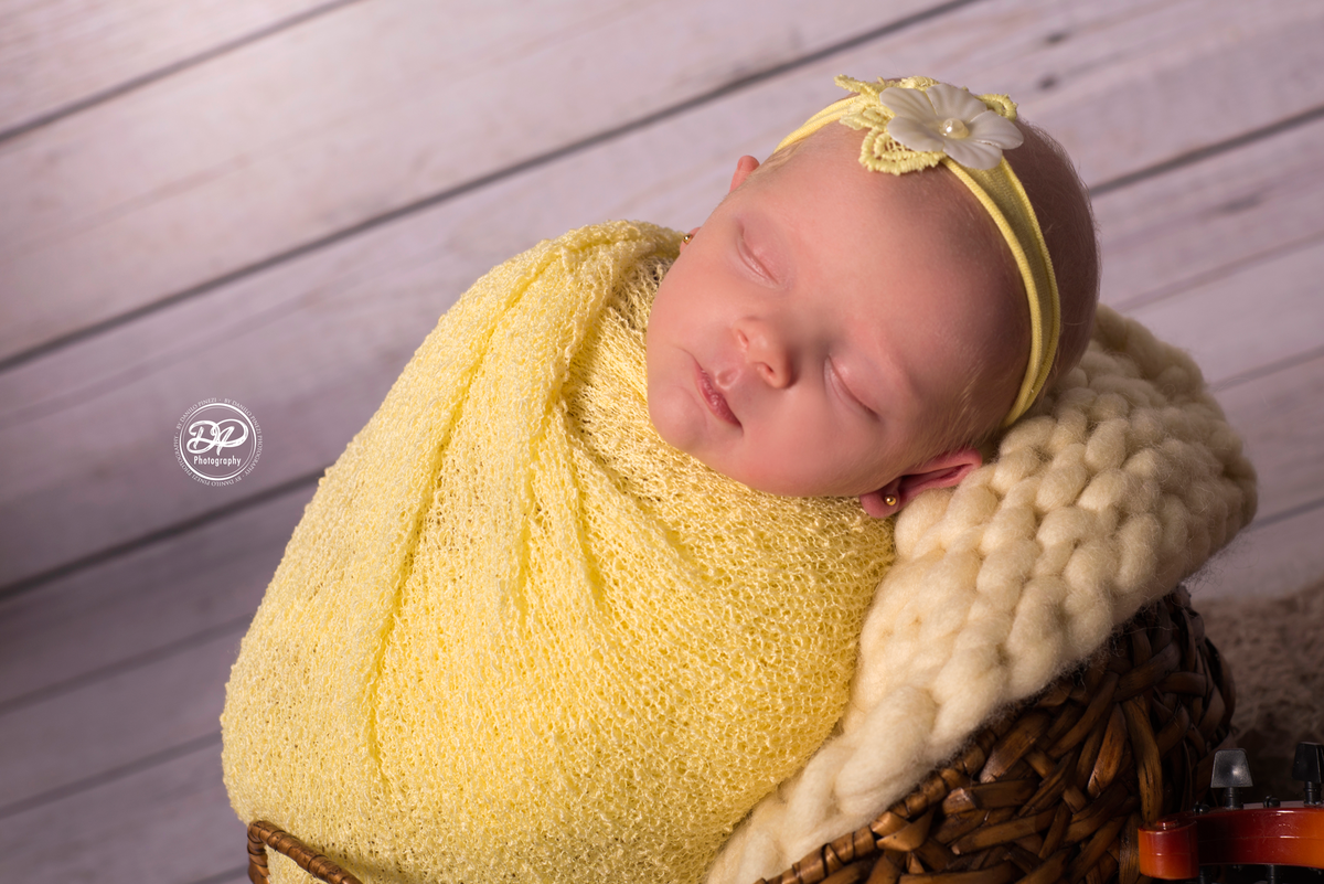 Ensaio newborn com a bebê enrolada no wrap amarelo posicionada dentro de um cesto, ornamentado com flores e um violino