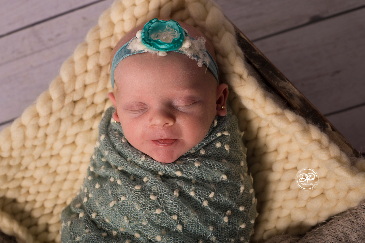 Ensaio newborn com a bebê enrolada no wrap verde pipoca posicionada dentro de um carrinho rústico, ornamentado com flores de cerejeira