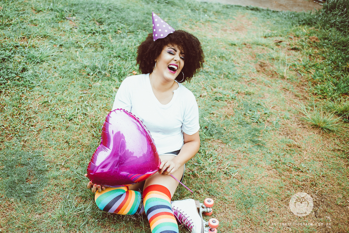 Ensaio feminino. Mulher com patins, balão metalizado de coração e chapéu de festa sentada na grama