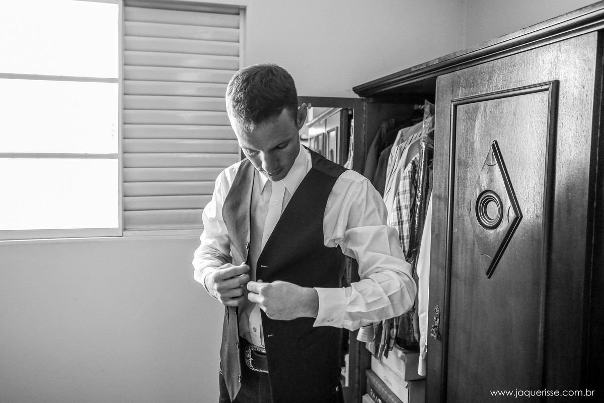 jaque risse, andre risse, estudio bebedouro, melhores fotografos, noivo abotoando o colete em frente ao guarda roupa com a luz vindo pela lateral de uma janela aberta durante o making of