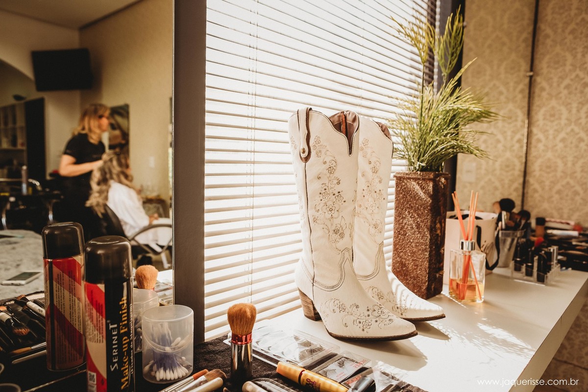 jaque risse, andre risse, estudio bebedouro, melhores fotografos, botas country da noiva sob a bancada com itens de maquiagem perto e uma linda luz vinda de uma janela com persianas