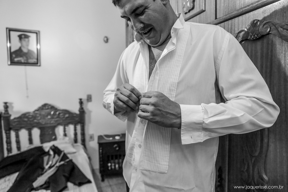 noivo sorridente abotoando a camisa em frente a seu guarda roupa e cama no click da fotógrafa de casamento Bebedouro SP Jaque Risse