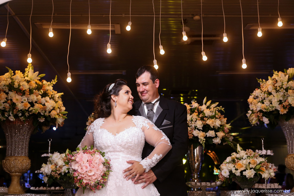noivos posando para foto em frente a mesa do bolo com lindas luzes penduradas ao fundo no click da fotógrafa de casamento Bebedouro SP Jaque Risse