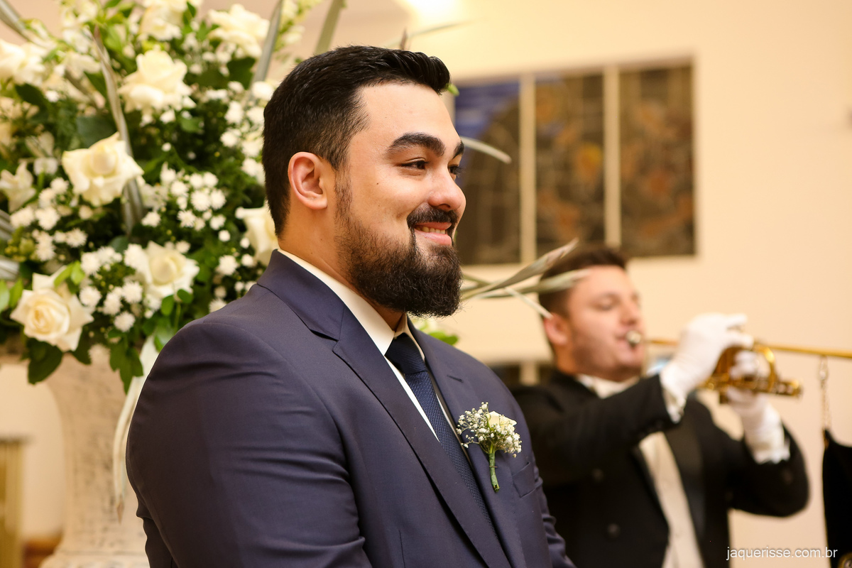 noivo no altar com o musico tocando trompete ao fundo aguardando a entrada da noiva pelo corredor da igreja registrado pelas lentas da fotografa jaque risse em bebedouro sp