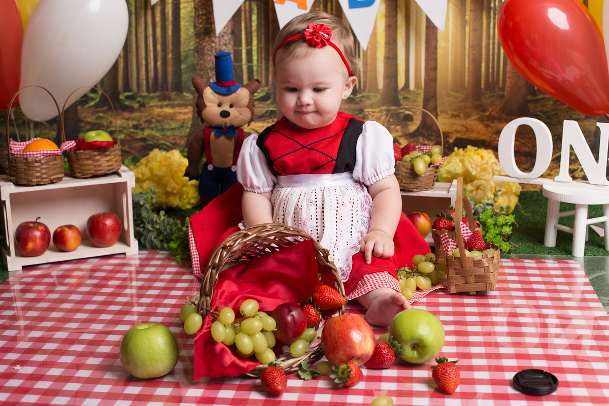 Foto de criança sentada na toalha xadrez. A bebê virou a cesta com frutas e ficou muito divertido essa imagem.
