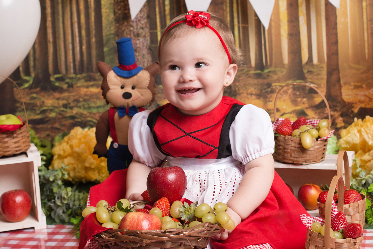 Book infantil de 1 ano da princesa Eloá. Ela está segurando uma cestinha de frutas naturais e está toda sorridente. 