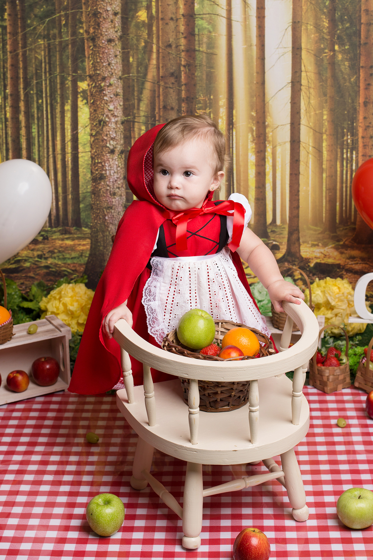 Foto da bebê no estúdio. Cenário com frutas. A bebê ficou de pé apoiada em uma linda cadeirinha retrô.