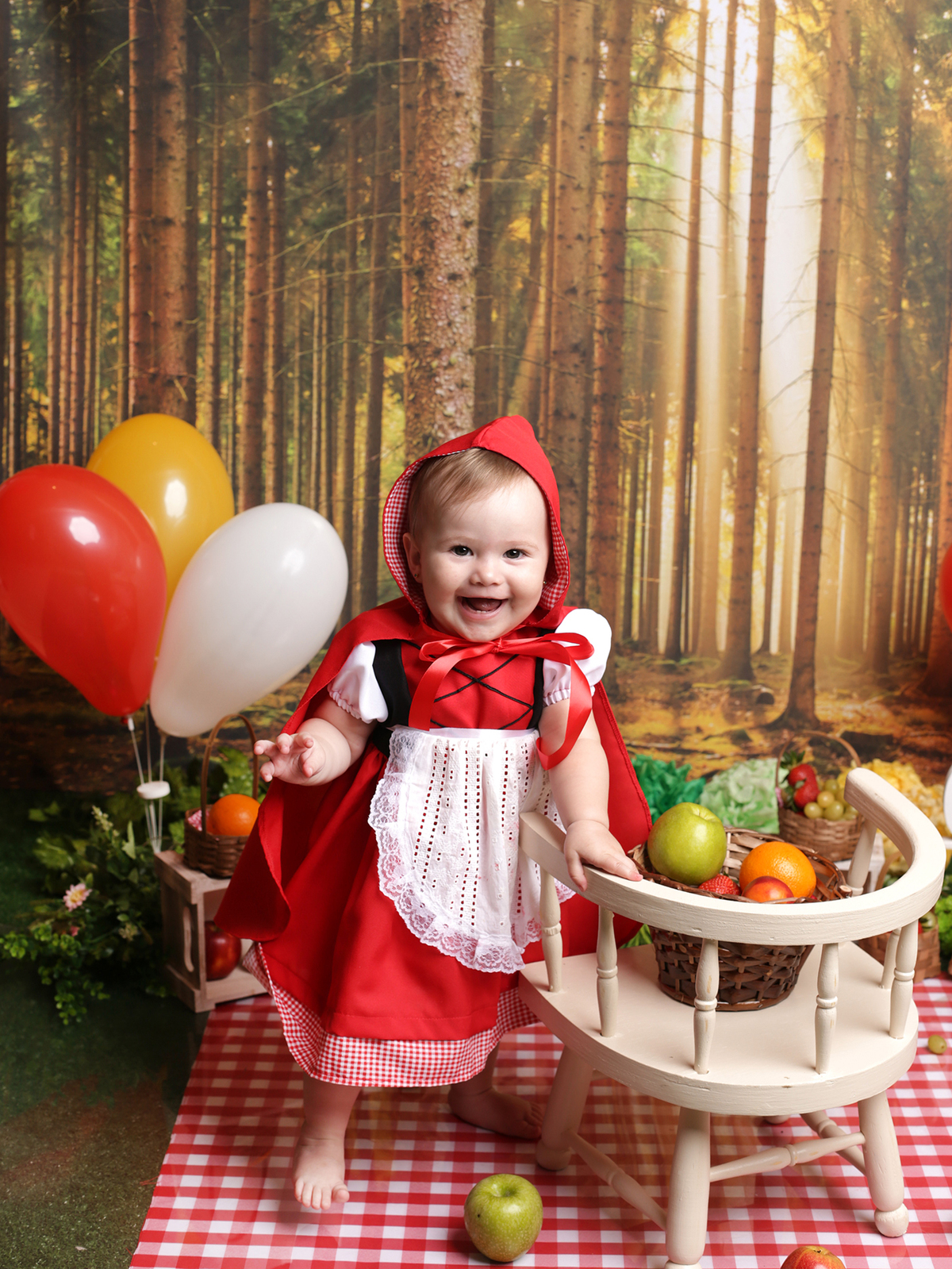 Ensaio no estúdio Edna Rosa Fotografia São Paulo SP.
 A bebê está usando um vestido vermelho com capa e capuz. O cenário floresta está mais radiante com os balões e frutas espalhadas nas cestinhas em vime.