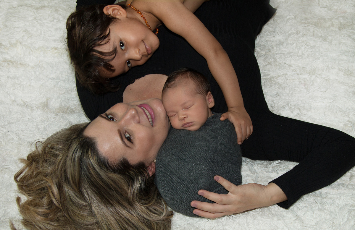 nsaio newborn com mãe e irmãozinho Theo. bebê enroladinho no wrap apoiado no ombro da mãe, irmão deitado e olhando pro bebê