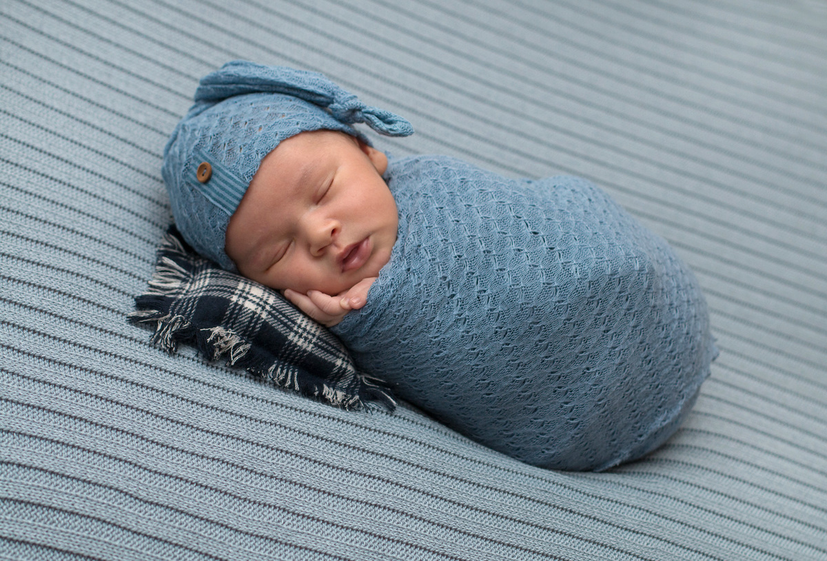 bebê enrrolado no wrep azul claro usando touca mesma cor. está deitado no puff sobre um travesseiro tecido xadrez