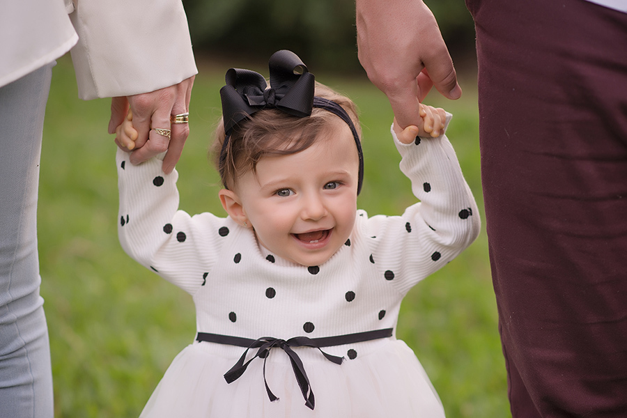 menina vestida de branco com bolinhas pretas segurando a mão dos pais