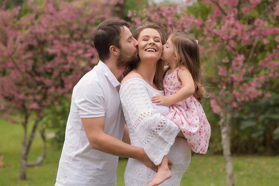 fotografia gestante em caxias do sul; Morgana Perini; Fotografia de família em Caxias do Sul; gestante ganhando beijo do marido e filha com árvores em flor