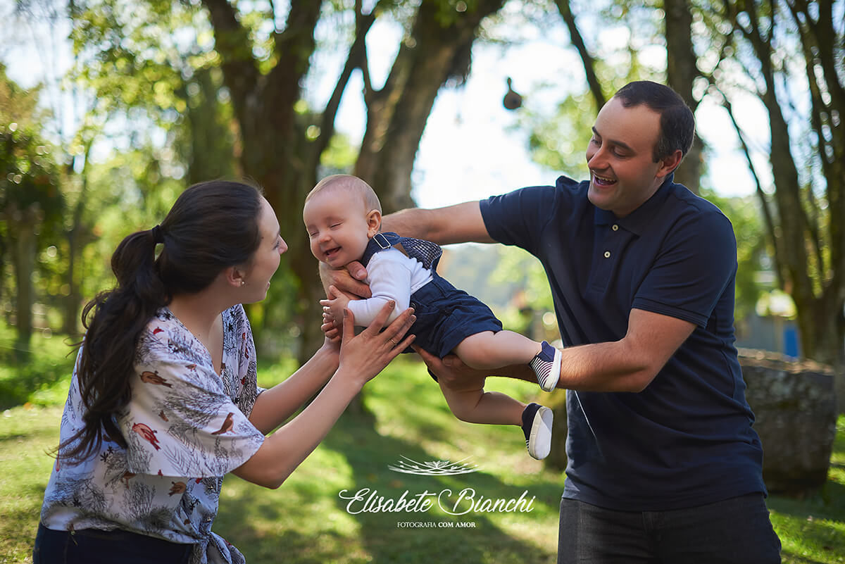 Pais de bebê de 6 meses brincando de aviãozinho com o filho, no ensaio de acompanhamento trimestral em São Marcos, RS.