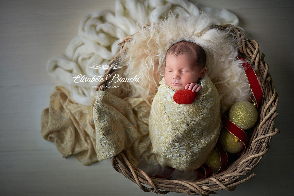 Bebê recém-nascida enrolada, dentro de cesto, segurando coração vermelho, em ensaio newborn pacotinho, Caxias do Sul, RS.