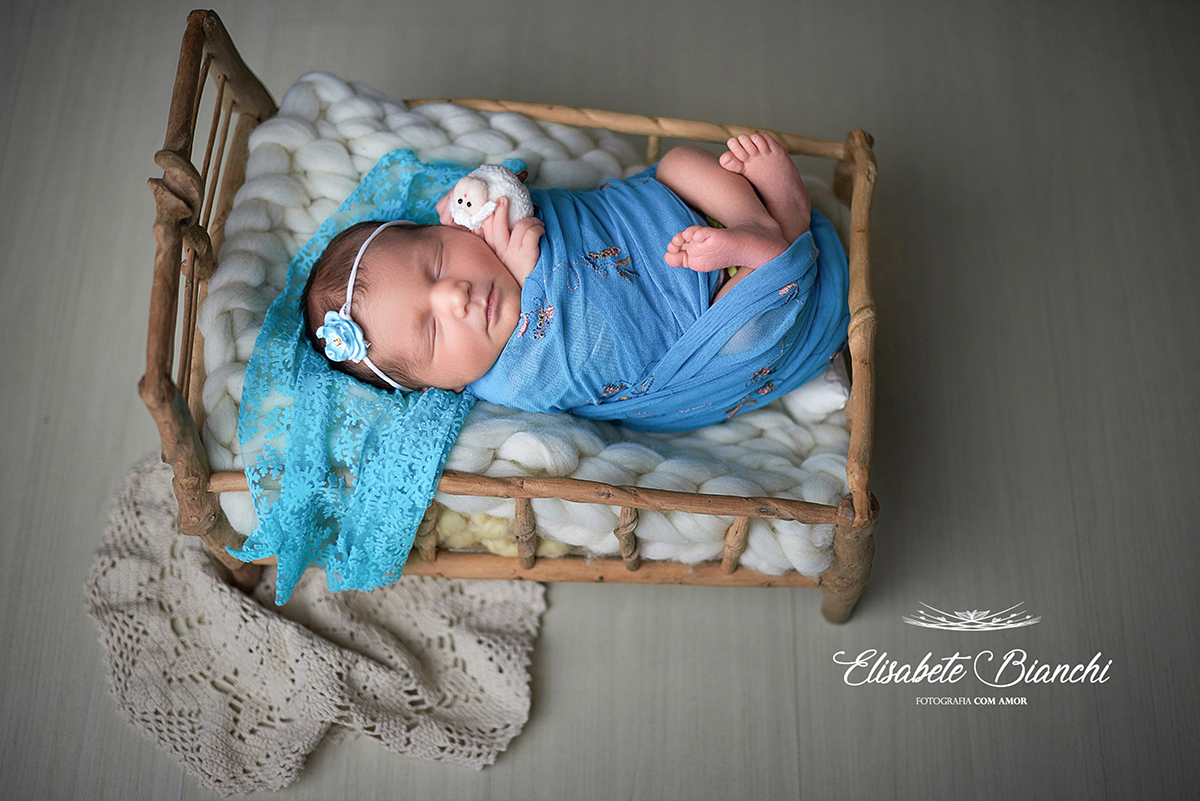 Recém-nascida enrolada em wrap azul, dentro da caminha e segurando ovelhinha, no ensaio newborn, em Caxias do Sul, RS.