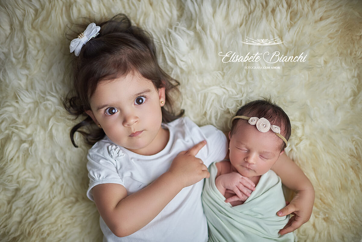 Menina deitada e abraçada na sua irmã caçula recém-nascida, no ensaio newborn, em Caxias do Sul, RS.
