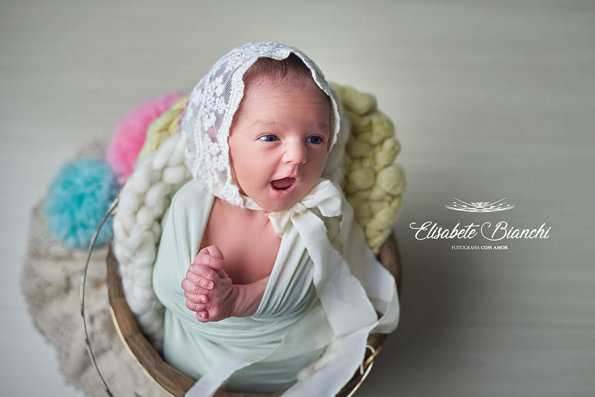 Bebê recém-nascida com mãos em oração, acordada no ensaio newborn, parecendo estar cantando, em Caxias do Sul, RS.