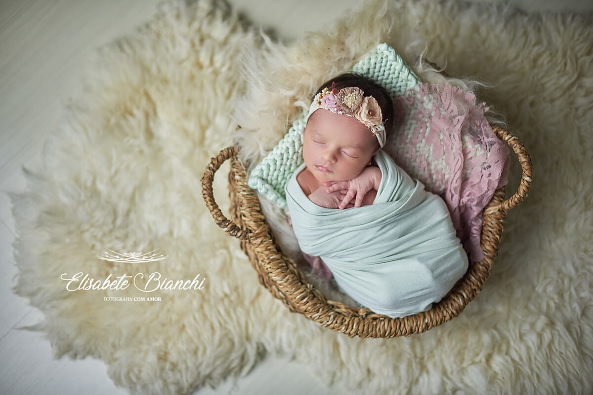 Recém-nascida enrolada, deitada em um cesto sobre um pelego, no ensaio newborn, em Caxias do Sul, RS.