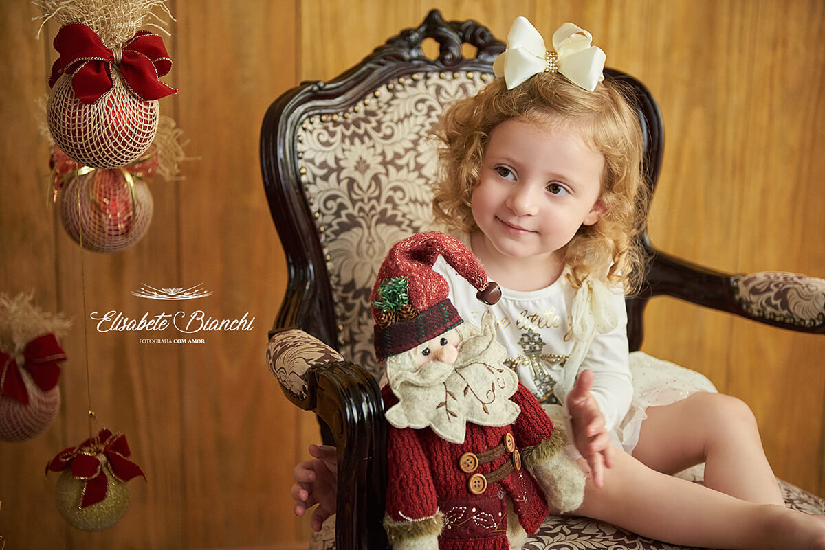 Menina down sentada em poltrona Luiz XV, segurando boneco de Papai Noel em ensaio natalino, em estúdio, Caxias do Sul - RS.