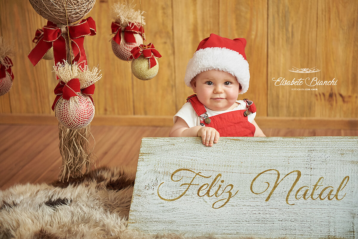Bebê com touca de Papai Noel, segurando placa de Feliz Natal, em ensaio de estúdio, Caxias do Sul - RS.