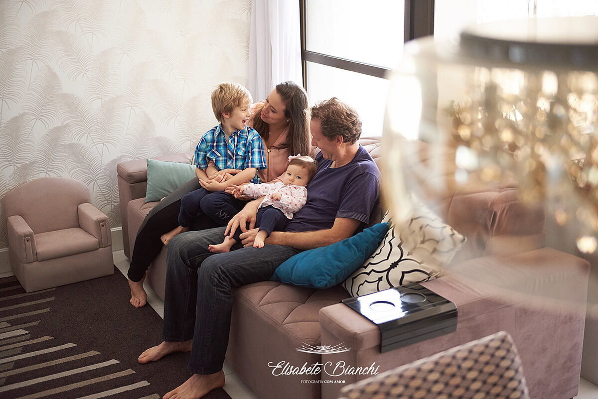 Família sentada no sofá da sala, com bebê de 7 meses no colo, em ensaio lifestyle na residência da família, em Caxias do Sul - RS.