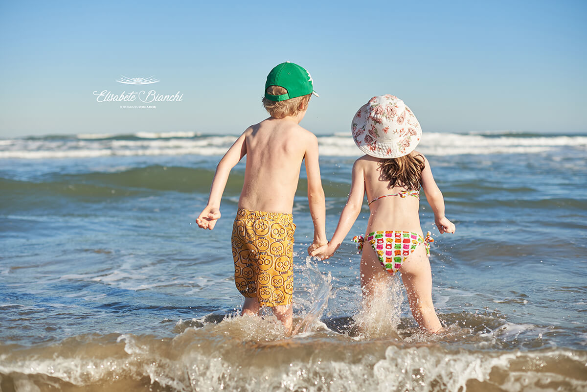 Crianças de mãos dadas entrando no mar de Bella Torres - SC, vistos de costas.