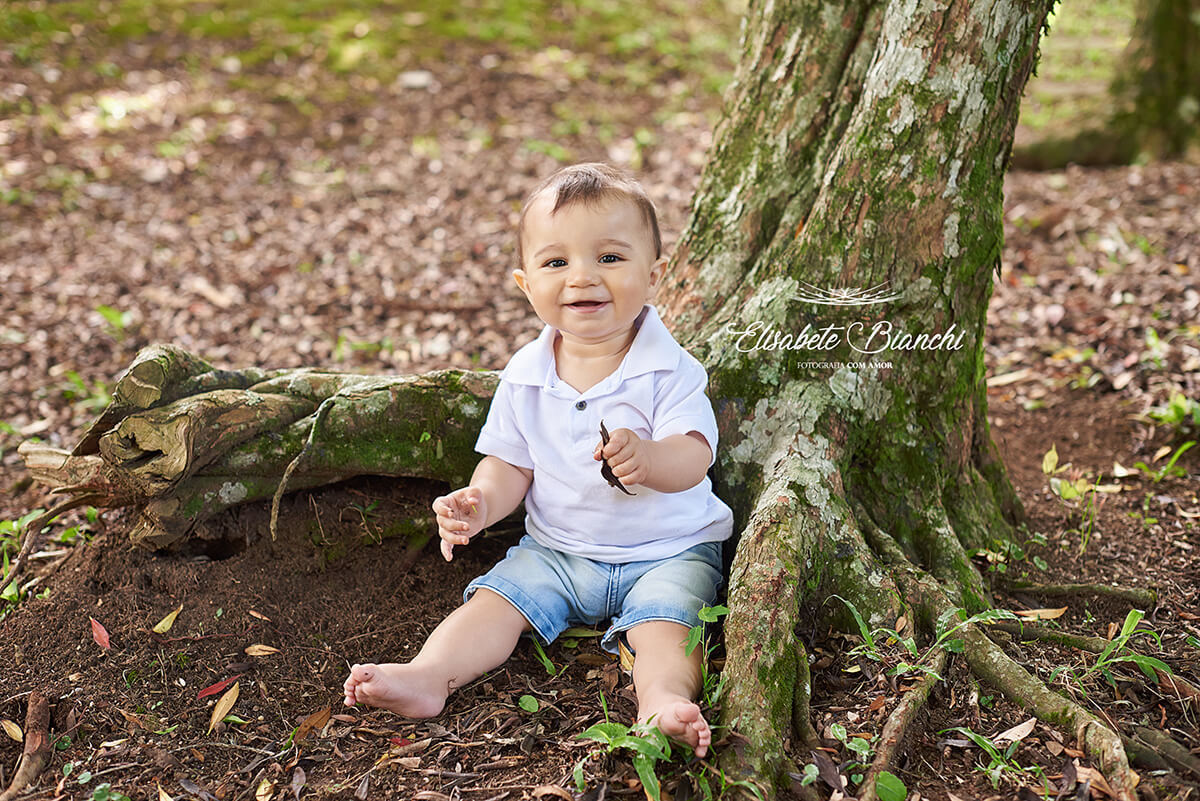 Bebê de 10 meses sorrindo, sentado sob uma árvore em ensaio fotográfico em Caxias do Sul - RS