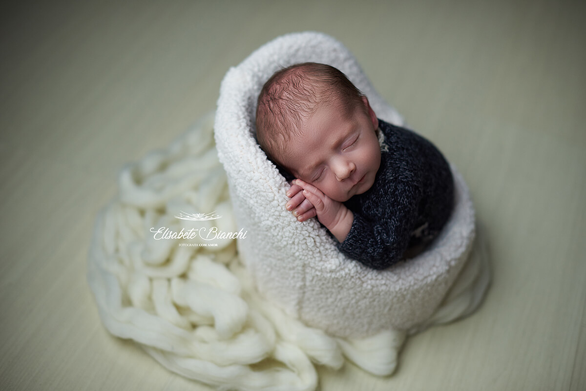 Bebê recém-nascido dormindo na poltrona, com as mãos apoiando o rosto, no ensaio Newborn, em Caxias do Sul - RS