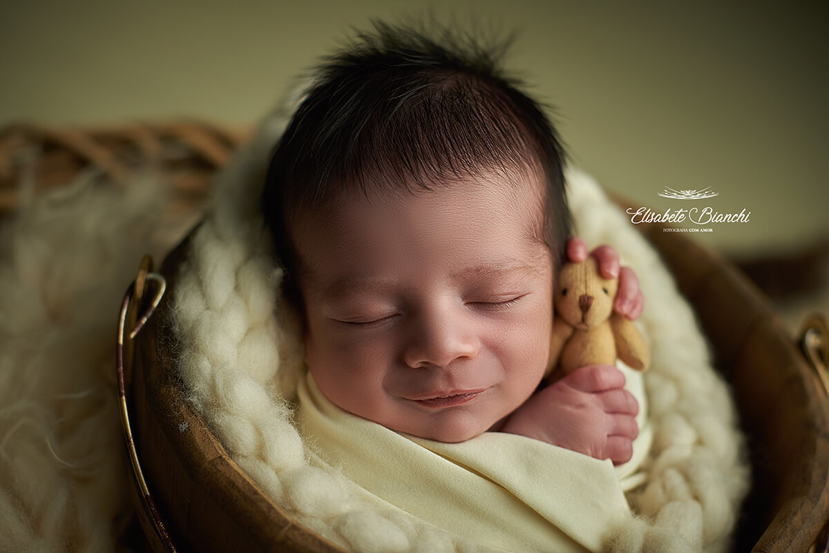 Bebê recém-nascido sorrindo, com um ursinho, e enroladinho, no ensaio newborn, em Caxias do Sul - RS.