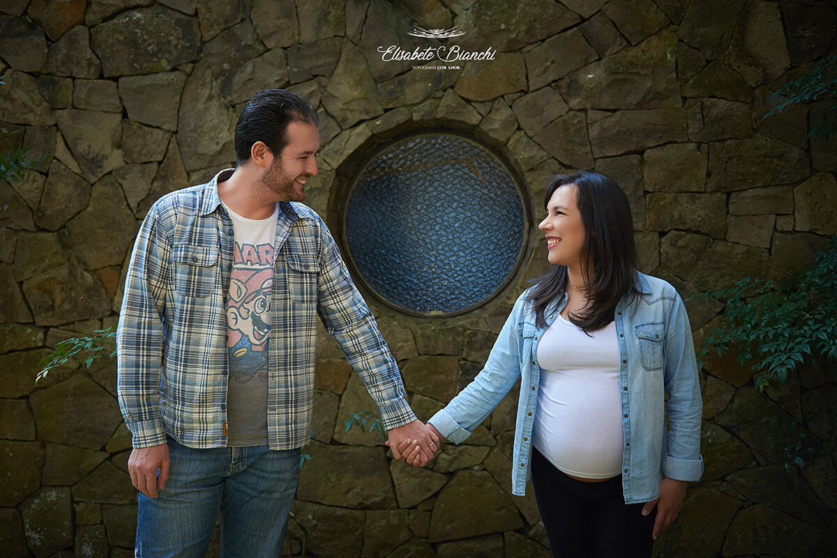 Casal gestante de mãos dadas, se olhando nos olhos e sorrindo, em frente à uma parede de pedras, com uma janela redonda, em ensaio fotográfico gestante, Caxias do Sul.