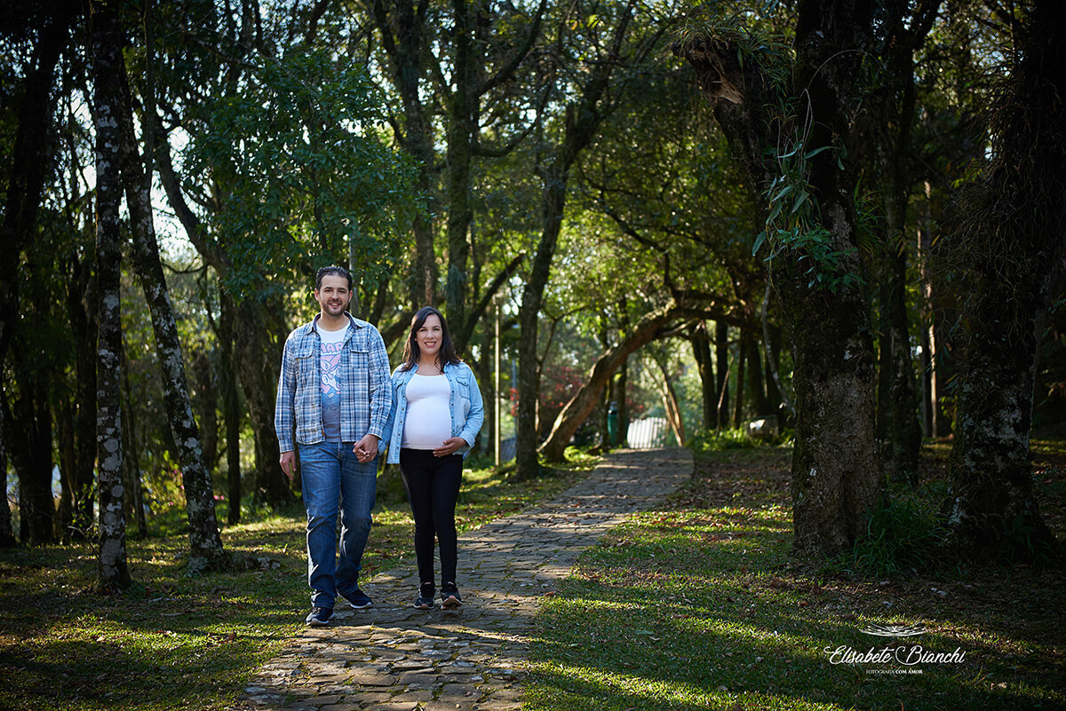 Casal de mãos dadas, caminhando em uma estradinha de pedras, em meio às árvores, em ensaio gestante externo, Caxias do Sul.