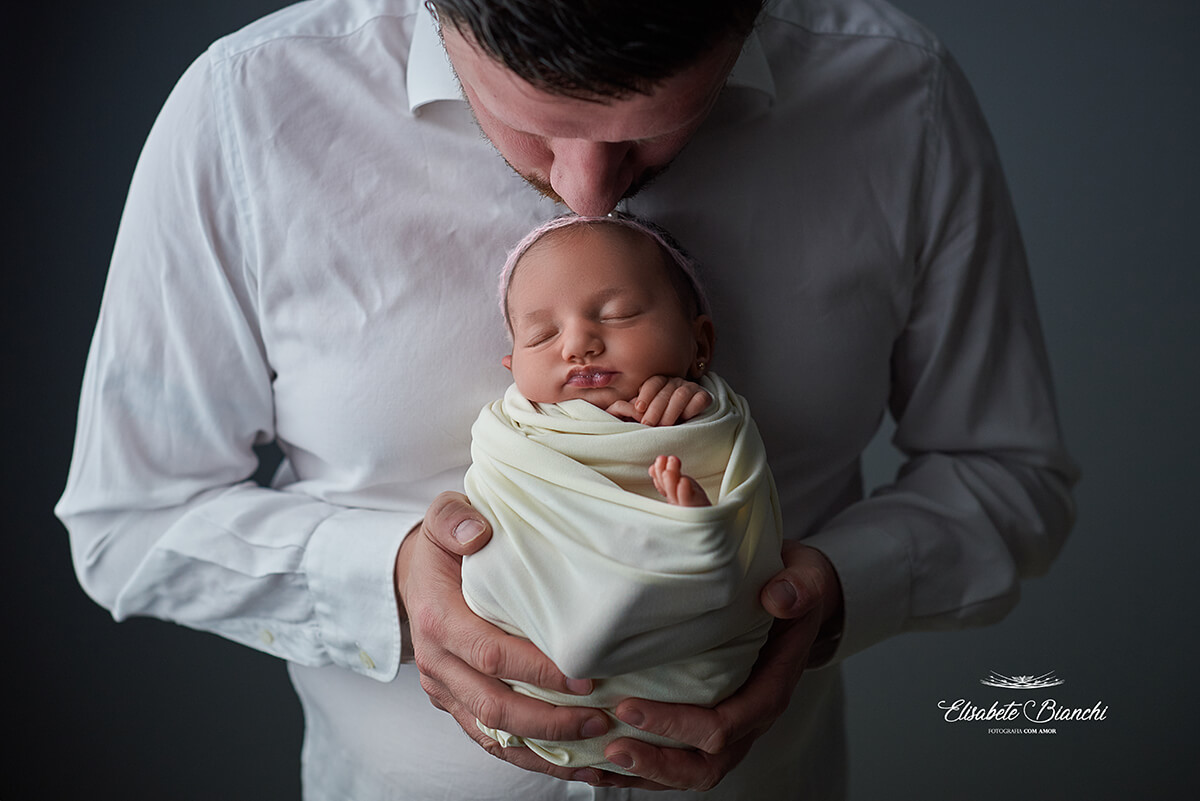 Pai segurando sua bebê recém-nascida sobre seu peito e beijando sua cabeça, em ensaio newborn, Caxias do Sul - RS