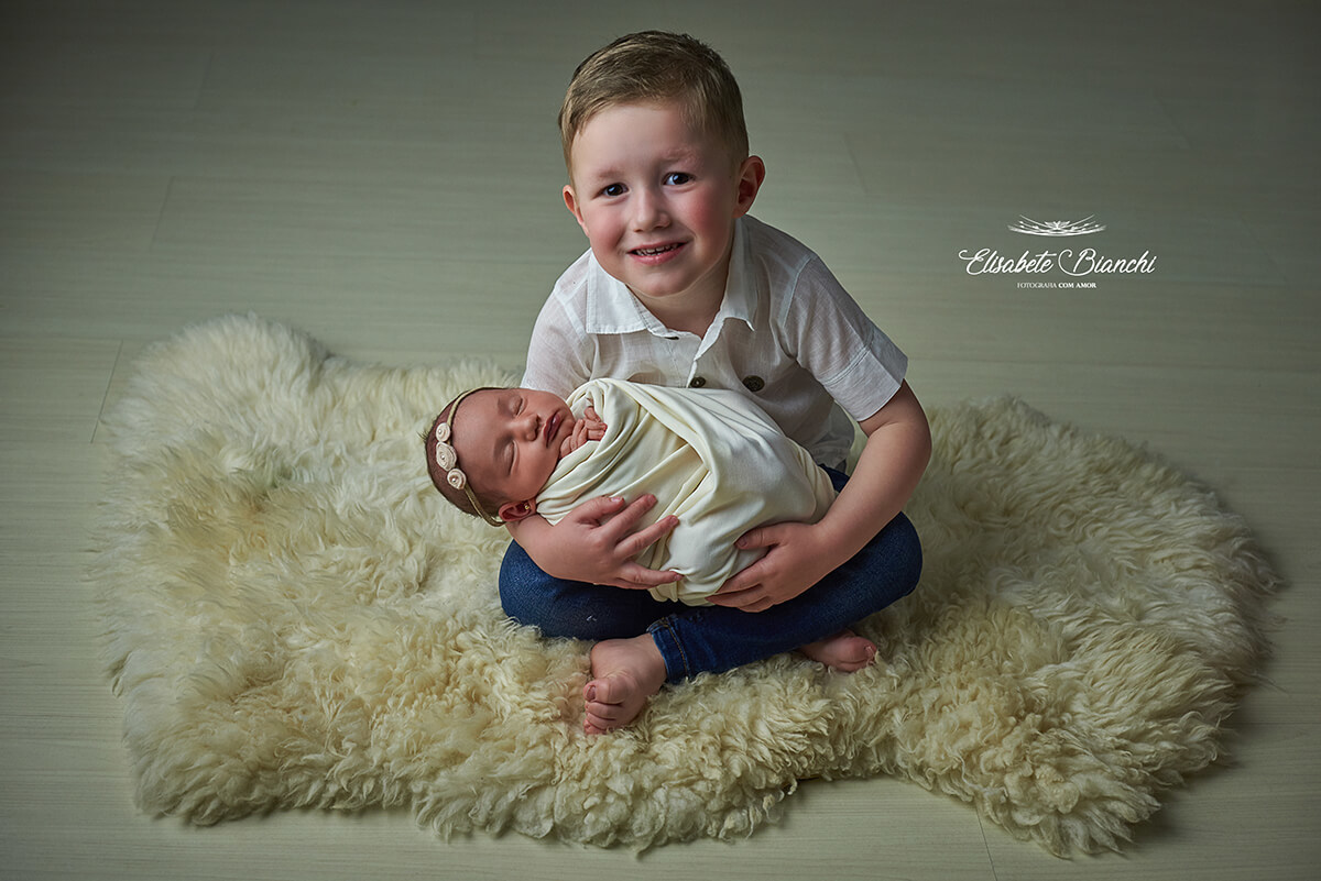 Irmão mais velho segurando no colo irmã recém-nascida, sentado com pernas de índio, sobre um pelego, em ensaio newborn, Caxias do Sul - RS