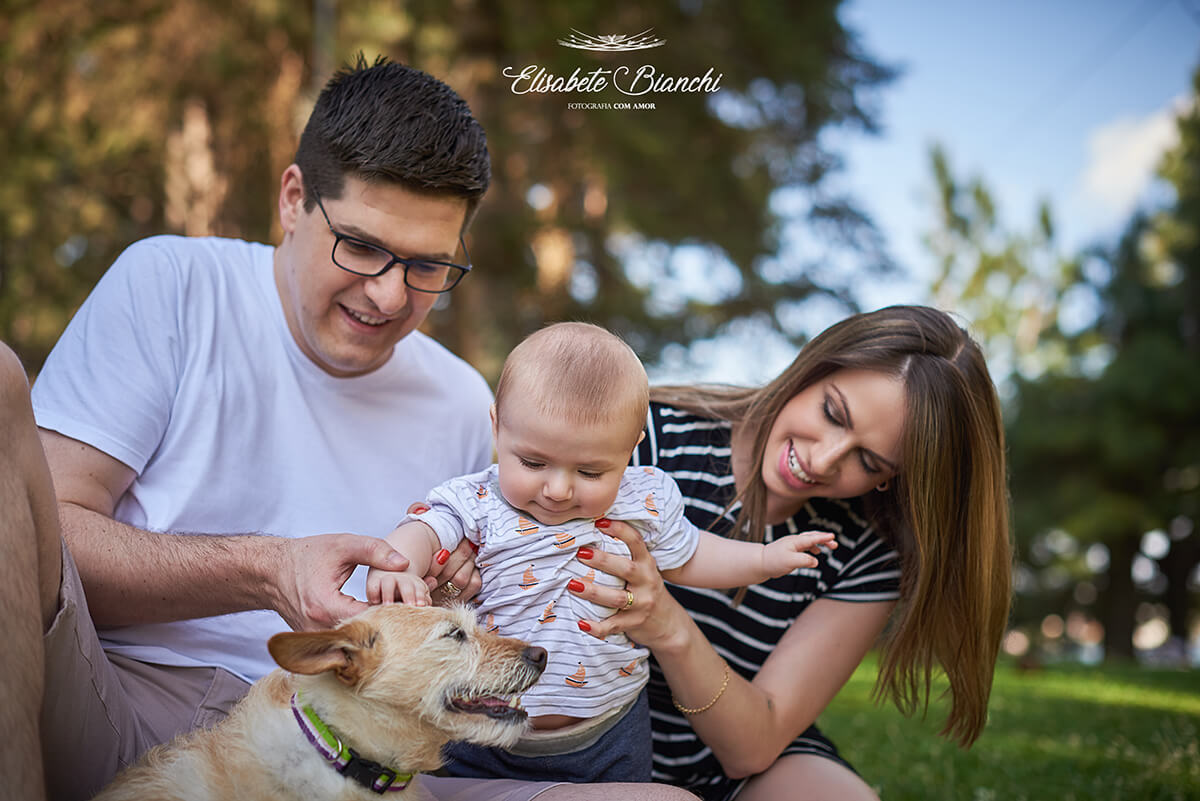 Família em um parque, com os pais ajudando o bebê a passar a mão na cabeça do cachorro.