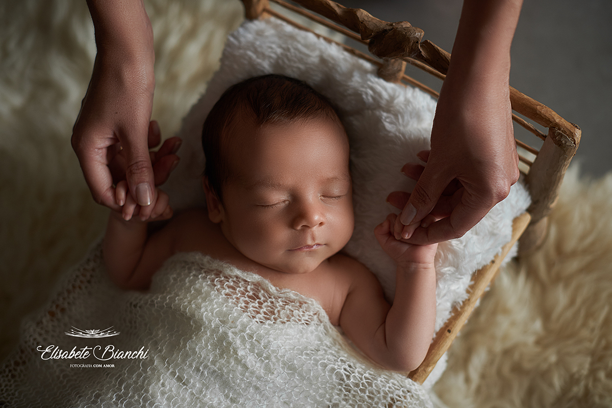 Bebê dormindo em uma caminha e segurando as mãos da mãe, em ensaio newborn.