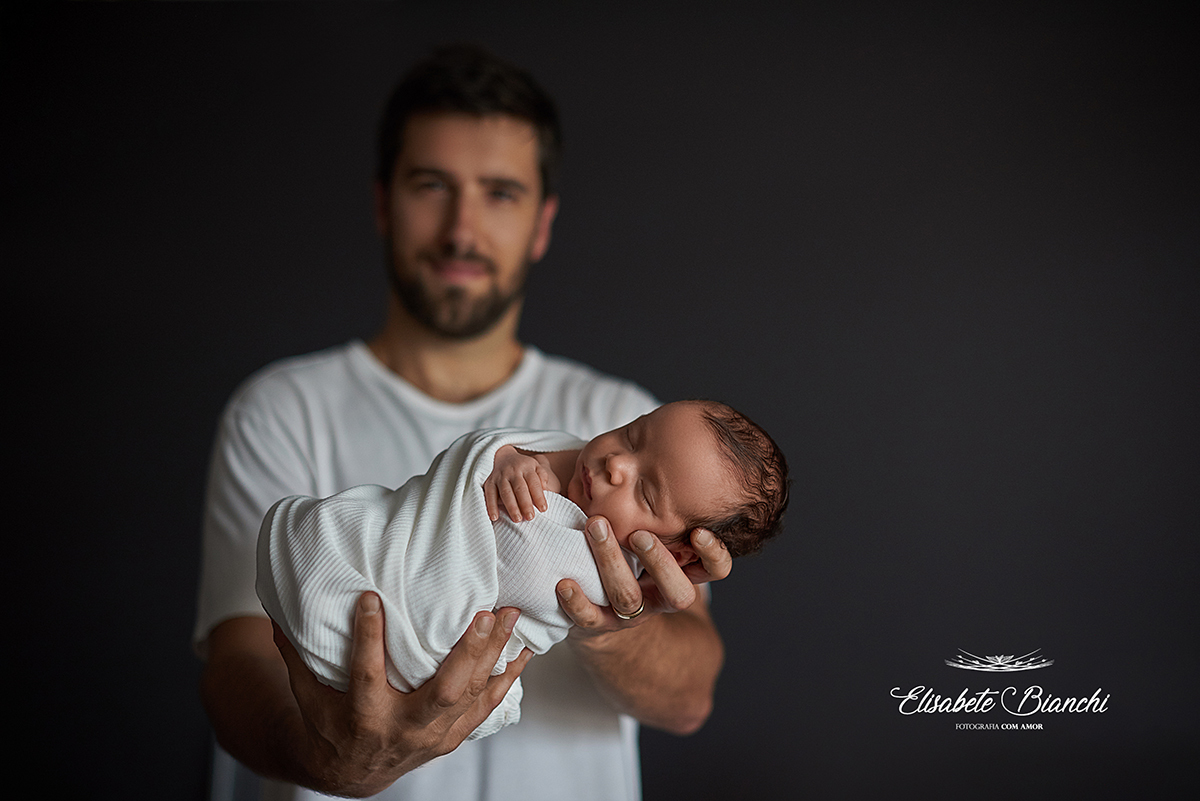 Bebê, enrolado em um tecido, nas mãos do pai, em ensaio newborn.