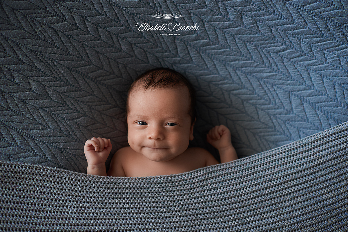 Bebê  nascido prematuro, olhando para a câmera, com os bracinhos para cima, debaixo do cobertor, em ensaio newborn.