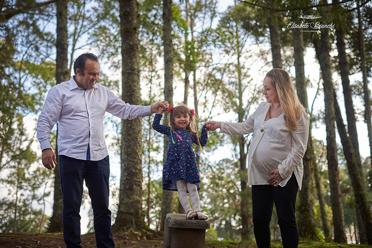Casal gestante ajudando filha mais velha a caminhar em um banco de parque, no ensaio fotográfico em Caxias do Sul.