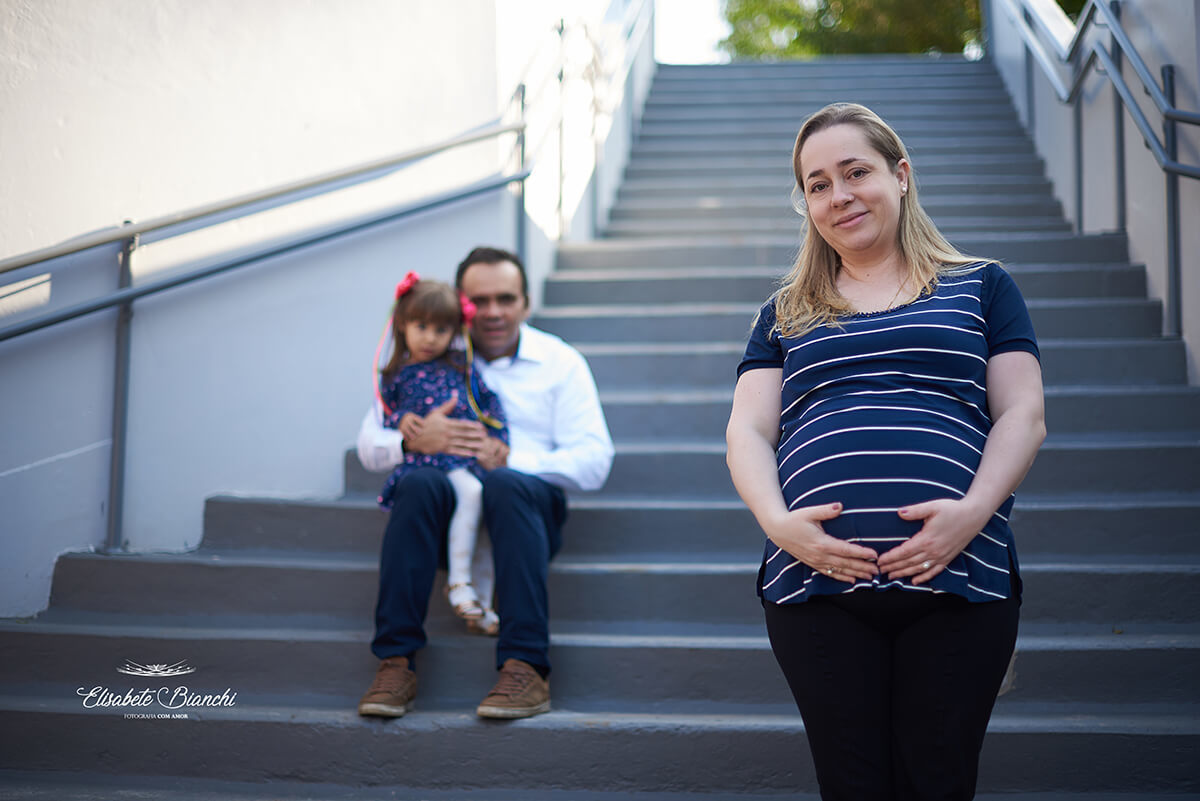 Família gestante, em escadaria. A mãe está de pé, a frente e em foco, e pai e filha, ao fundo, sentados, fora de foco, em ensaio fotográfico Caxias do Sul.